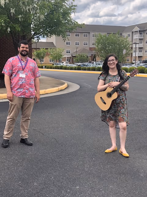 A man and a woman standing outside on a paved area near a large residential building. The man is wearing a pink patterned shirt and beige pants, and the woman is wearing a floral dress and holding an acoustic guitar. Trees and parked cars are visible in the background under a partly cloudy sky.
