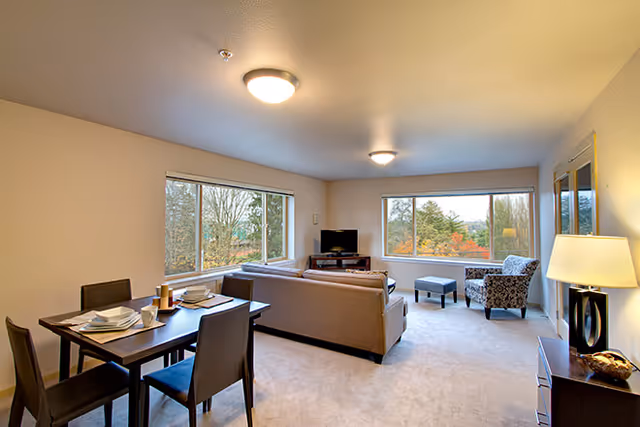A bright living room and dining area with large windows showing trees outside. The room features a dining table set for four, a beige sofa facing a TV on a stand, a patterned armchair with an ottoman, and a floor lamp on a side table.