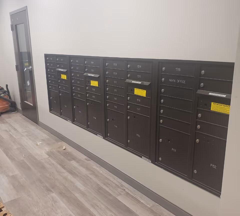A hallway with a row of black mailboxes mounted on a white wall. The mailboxes are labeled with numbers and some have yellow notices attached. There is a door with a glass panel at the end of the hallway and a vacuum cleaner is partially visible on the left side. The floor has light-colored wood or laminate flooring.