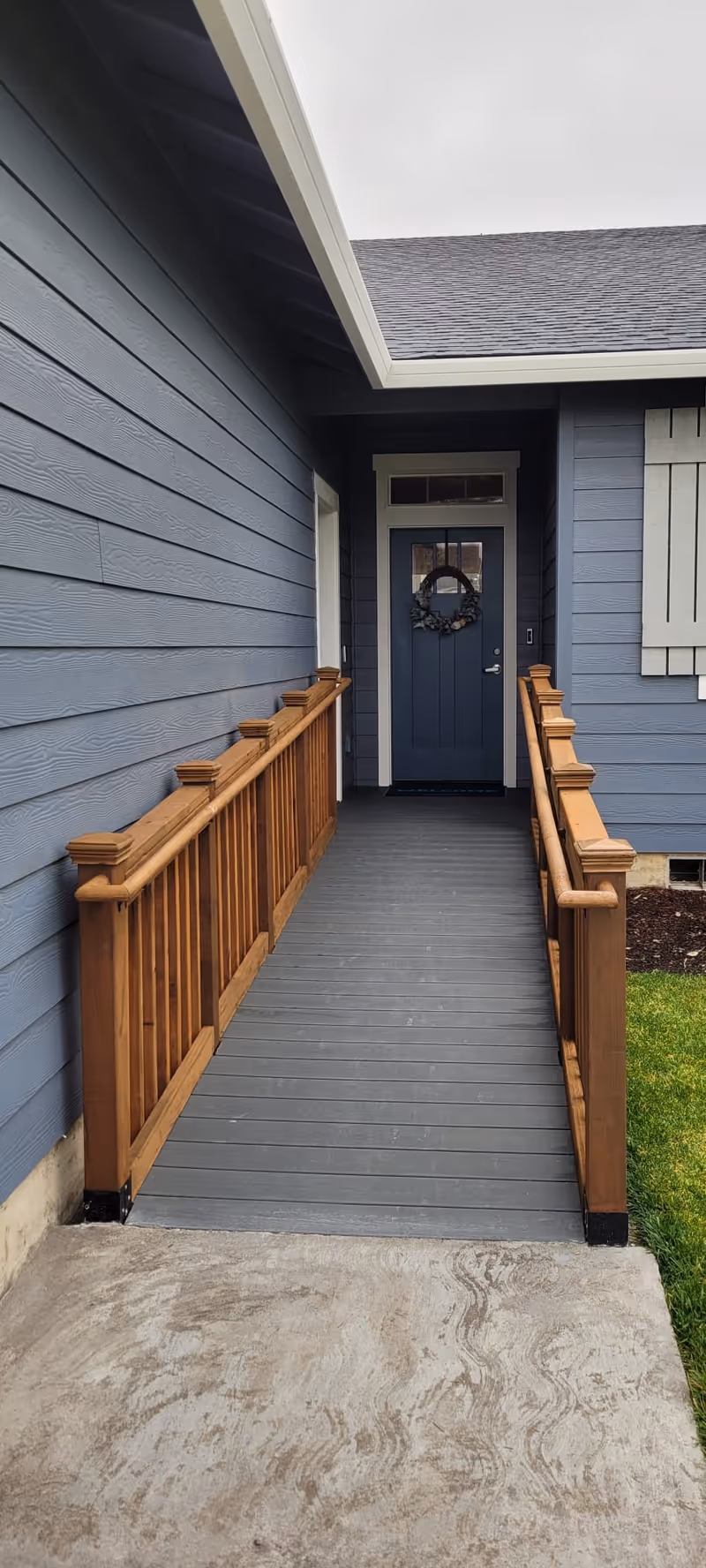 A wooden ramp with handrails on both sides leading to a blue front door of a house with gray siding. The door has a decorative wreath hanging on it. There is a small patch of grass visible to the right of the ramp.