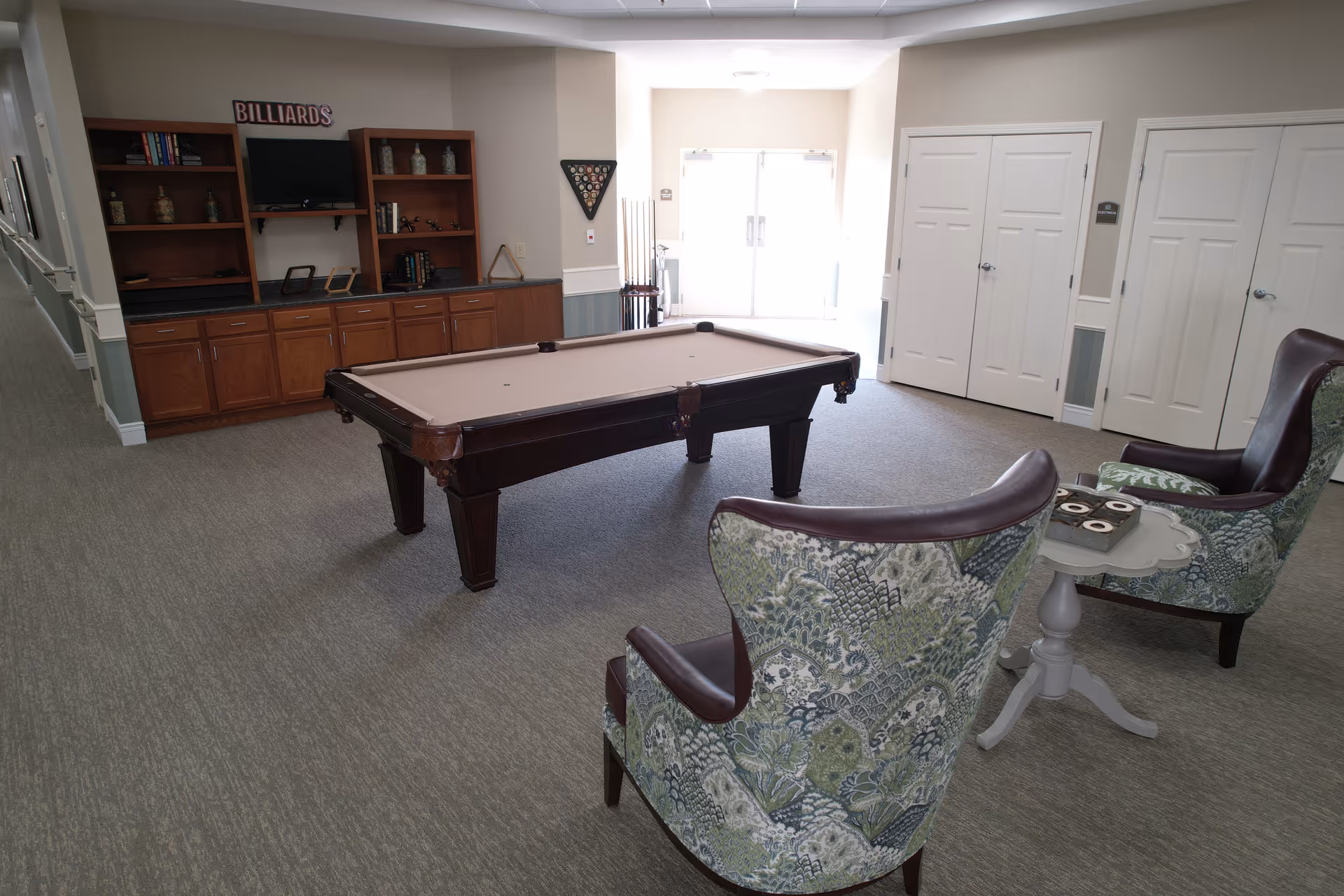 A senior living facility game room featuring a billiards table in the center, two patterned armchairs with leather trim facing each other beside a small white table, and built-in wooden shelves and cabinets along the back wall. The room has carpeted flooring, white double doors, and a bright entrance with glass doors in the background.