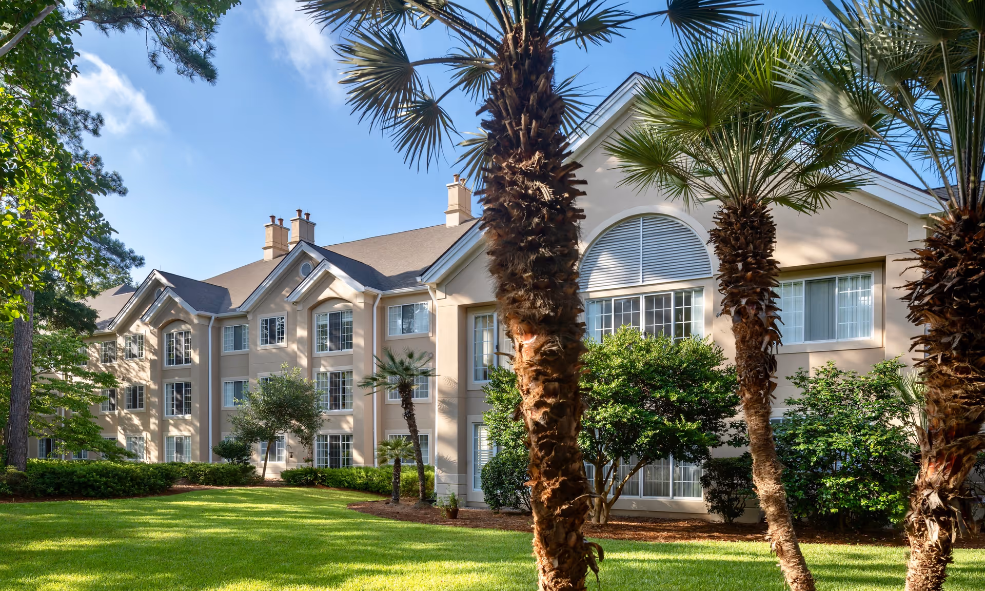 Exterior front view of a multi-story residential building with palm trees, shrubs, and a manicured lawn under a blue sky.