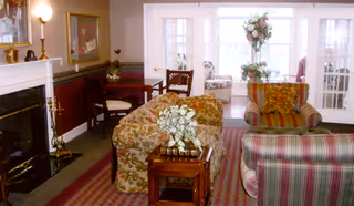 A cozy living room with floral and striped upholstered sofas and armchairs arranged around a wooden coffee table with a flower arrangement. There is a fireplace on the left side with a lamp and decorative items on the mantel. In the background, there are glass-paneled double doors leading to a sunlit room with more seating and a tall floral arrangement.