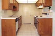 A compact kitchen with wooden cabinets on both sides, a white countertop, a dishwasher on the left, and a stove with an oven on the right. The floor is covered with light-colored tiles, and there is a closed door at the far end of the kitchen.