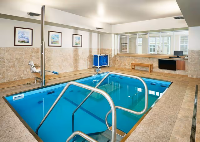 Indoor therapy pool with stainless steel handrails and steps leading into the water, surrounded by beige tiled flooring and walls. There is a white chair lift on the left side for accessibility, three framed paintings on the wall, a blue laundry cart, a wooden bench, and a desk with a computer in the background.