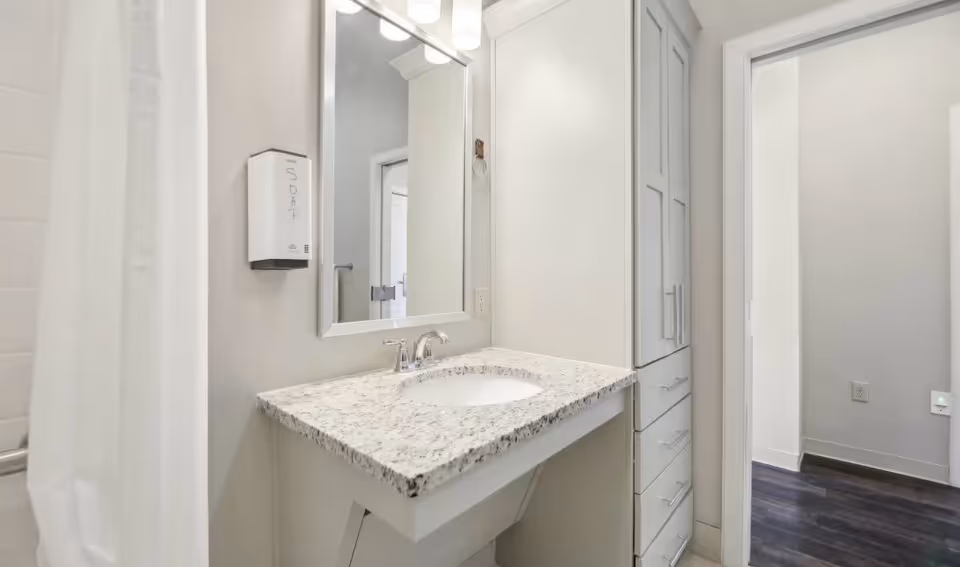 Bright modern bathroom with a granite-topped vanity, undermount sink, mirror, tall storage cabinet and a shower curtain visible at left.