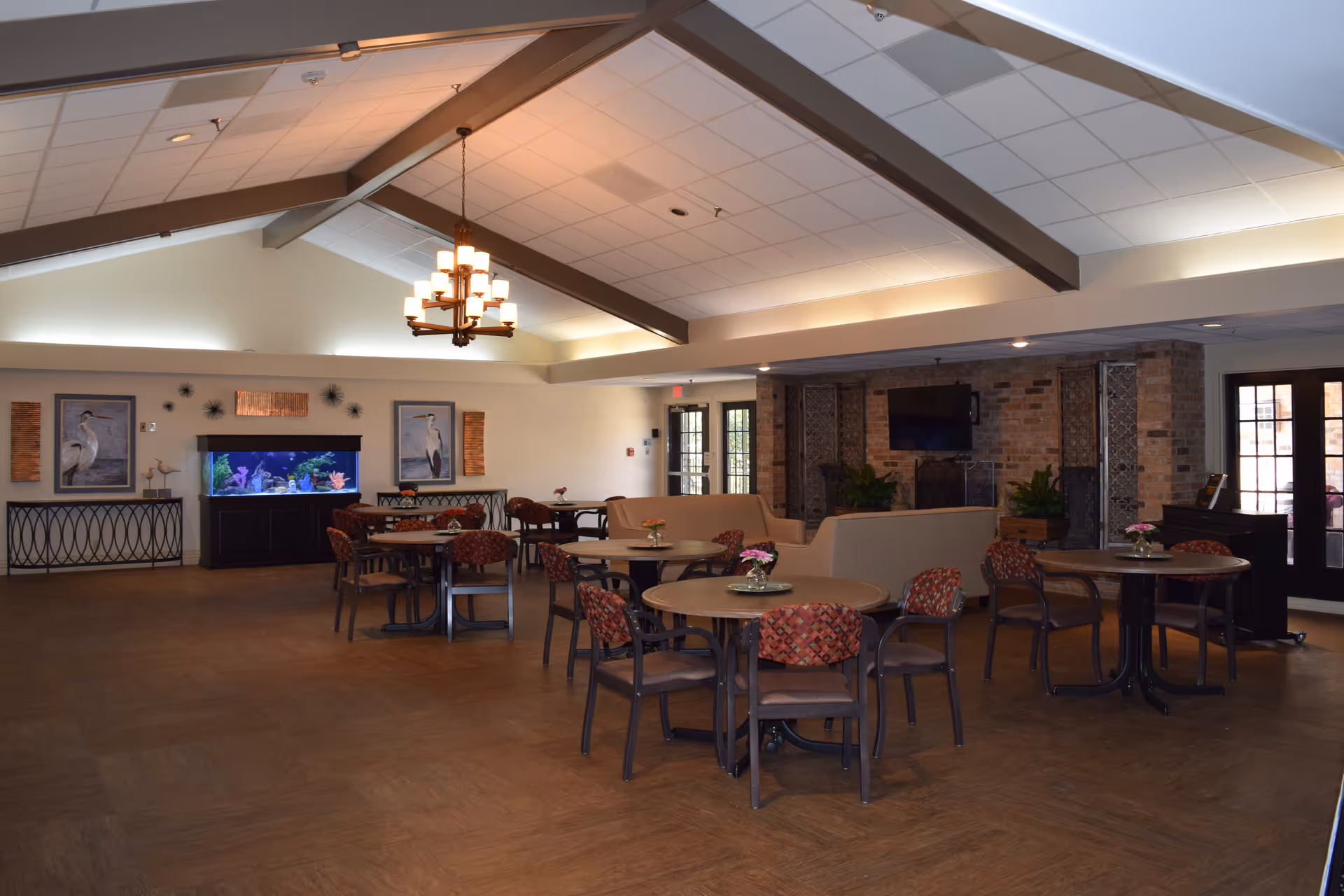 A spacious common area with round tables and chairs arranged for seating. The room features a high ceiling with exposed beams and a chandelier. There is a large fish tank against one wall, flanked by framed bird artwork. A beige sofa set is positioned near a brick wall with a mounted flat-screen TV and a fireplace. Double glass doors lead outside, allowing natural light to enter the room.