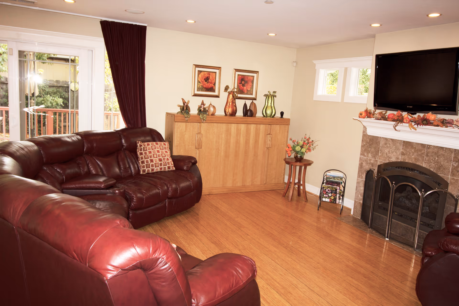 A cozy living room with a large burgundy leather sectional sofa, a wooden floor, and a fireplace with a mounted flat-screen TV above it. The fireplace mantel is decorated with autumn-themed garlands. There is a wooden cabinet against the wall with decorative vases and framed floral artwork above it. A small round table with a flower arrangement and a magazine rack are also visible. Sliding glass doors with dark curtains lead to an outdoor area.
