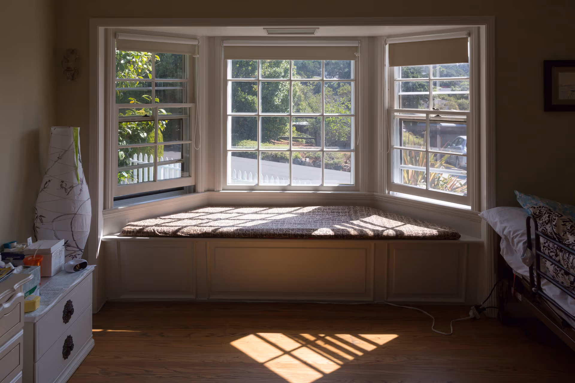 A cozy window seat with a cushion in a bay window area, sunlight casting shadows on the wooden floor. To the left is a white dresser with various items on top and a tall decorative lamp. To the right is part of a bed with patterned bedding.