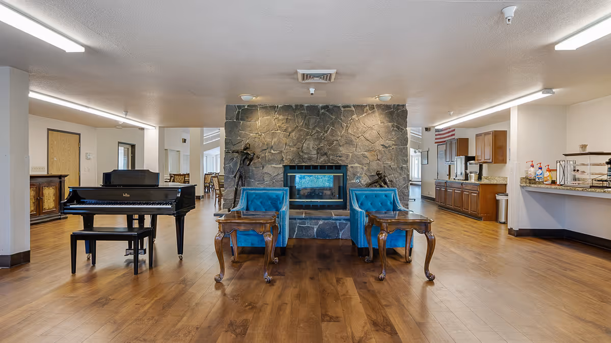 Spacious senior living common area with a stone fireplace flanked by two blue armchairs, a grand piano to the left, and a kitchenette to the right.