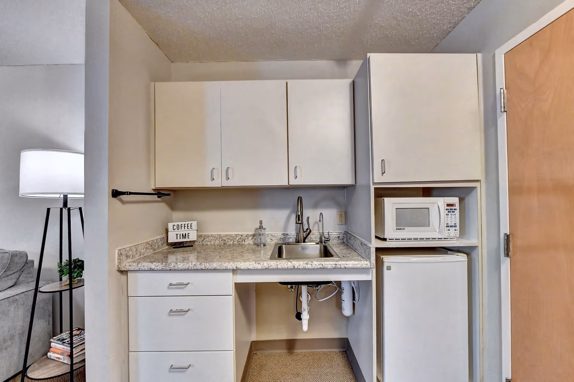 Small kitchenette with a sink, upper and lower cabinets, a microwave and mini refrigerator, and a countertop sign reading "COFFEE TIME".