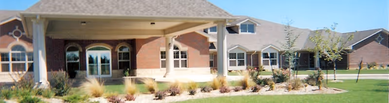 Exterior view of a single-story brick building with multiple windows and a covered entrance, surrounded by landscaped grass, plants, and small trees under a clear sky.