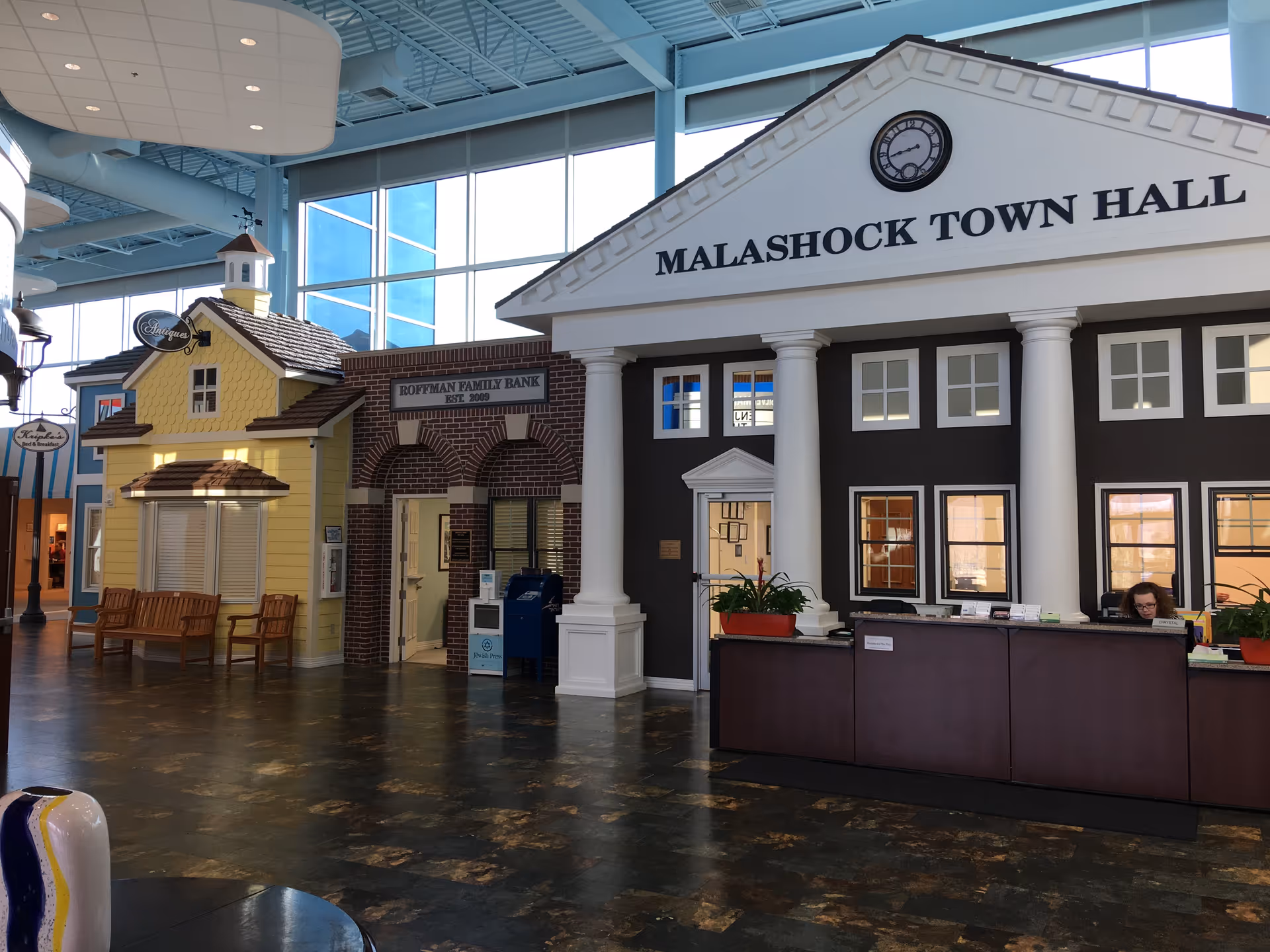 Interior commons area featuring a mock 'Malashock Town Hall' facade with columns, a reception desk, and adjoining small storefront-style rooms under a high ceiling.