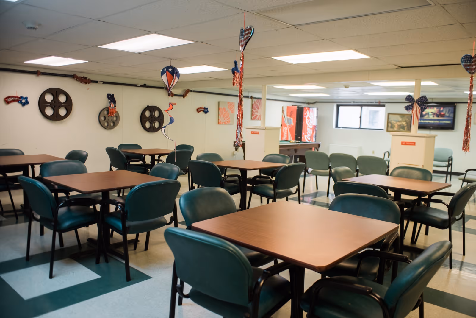 A common area with multiple square wooden tables surrounded by green cushioned chairs. The room has a low ceiling with fluorescent lights and patriotic decorations hanging from the ceiling and walls. There is a Coca-Cola vending machine and a pool table in the background, along with a television mounted on the wall.