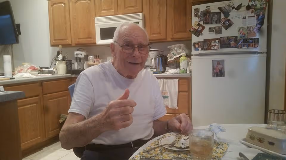 An elderly man wearing glasses and a white t-shirt is sitting at a kitchen table giving a thumbs-up gesture. The kitchen has wooden cabinets, a microwave, a refrigerator covered with photos and magnets, and various kitchen items on the counters. On the table in front of him is a plate with a dessert and a glass of beverage.