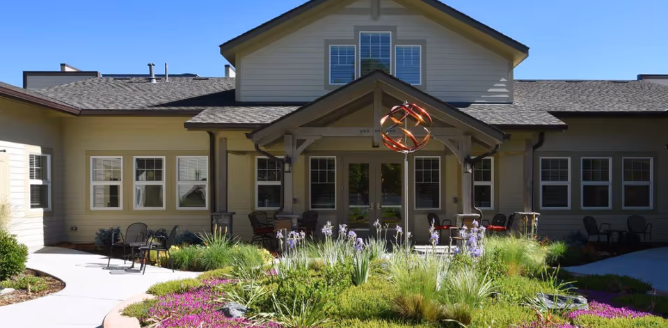 Outdoor view of a senior living facility named Gardens at Columbine showing a landscaped garden with purple flowers and green shrubs in front of a building with beige siding and multiple windows. There is a covered patio area with chairs and a decorative metal sculpture in the center of the garden.