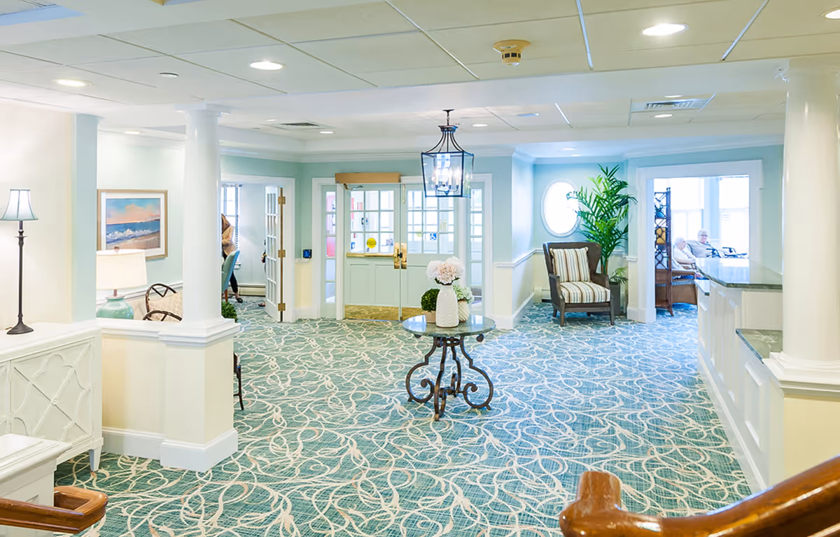 A bright and spacious common area in a senior living facility with patterned carpet, white pillars, a round table with flower vases in the center, a striped armchair near a potted plant, and large windows letting in natural light. Two elderly people are seated in a room visible through an open doorway on the right.