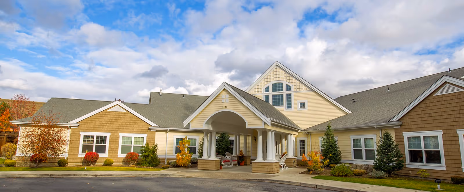 Exterior front view of a single-story senior living facility building with a covered entrance supported by white columns. The building has a combination of beige and light yellow siding with white trim, surrounded by small shrubs and trees under a partly cloudy blue sky.