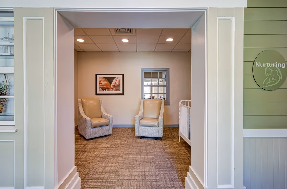 A small interior sitting room seen through a wide doorway with two beige armchairs, framed artwork, and a windowed wall.