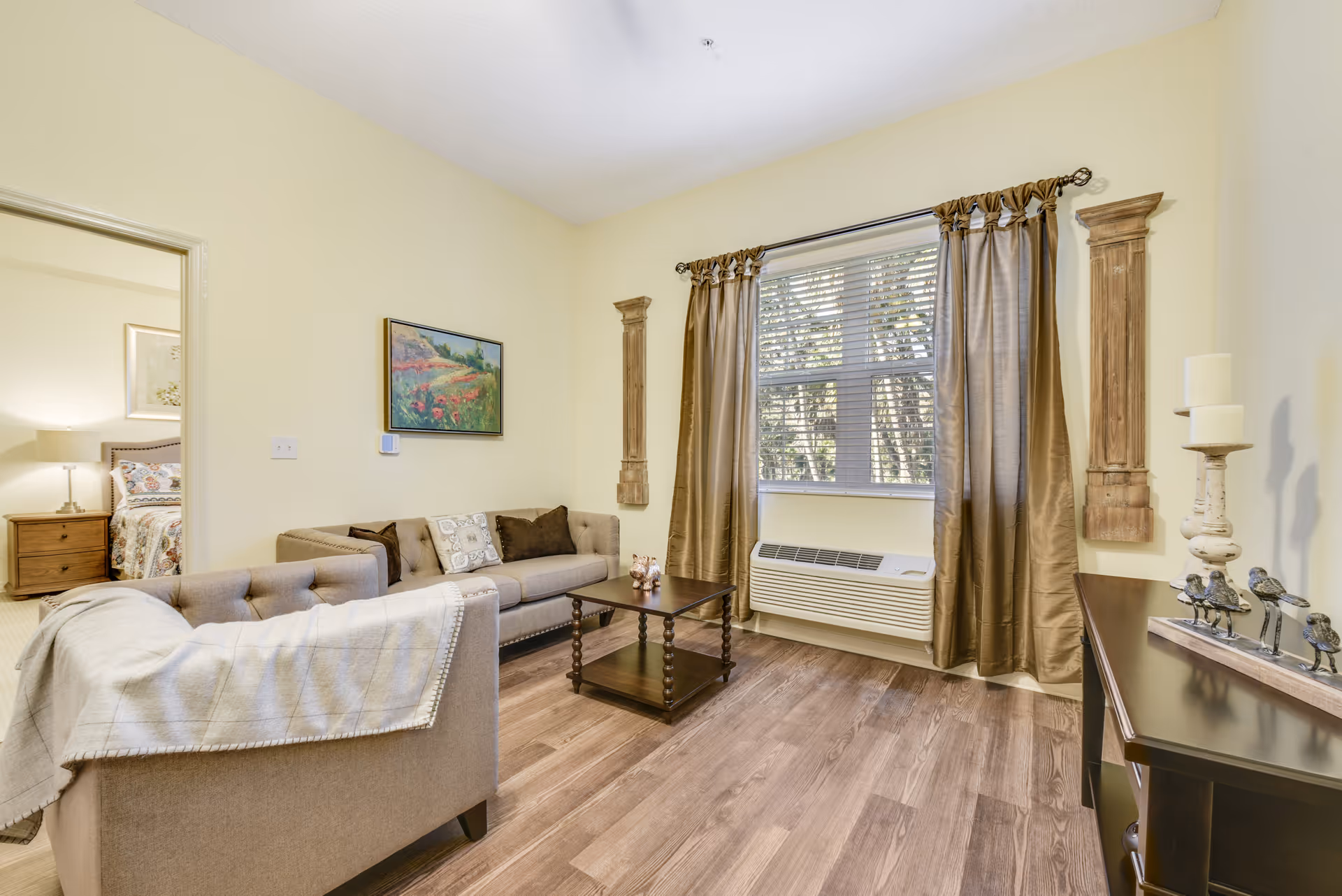 A cozy living room with beige walls and wood flooring featuring a beige sofa and armchair with a throw blanket. A wooden coffee table sits in the center. The window has brown curtains and blinds, with an air conditioning unit below. Decorative wall art and candle holders adorn the walls and furniture. A bedroom with a bed and nightstand is visible through an open doorway.