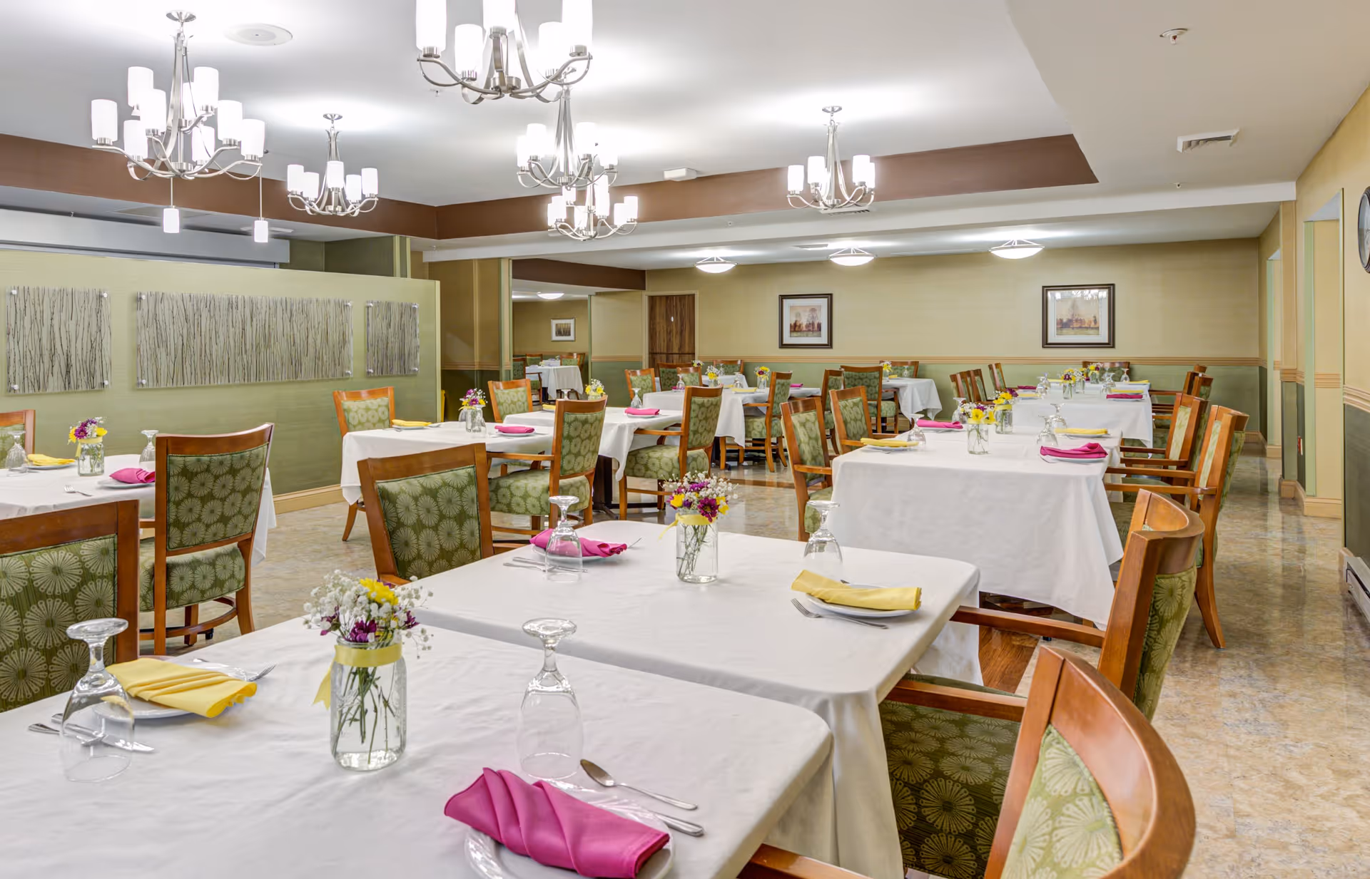 Spacious dining room with multiple tables set with white tablecloths, colorful napkins, flower centerpieces, and chandeliers.