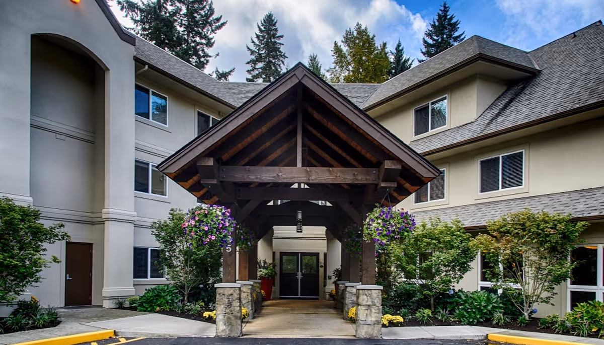Entrance of a senior living facility with a wooden covered walkway supported by stone pillars, surrounded by greenery and hanging flower baskets, under a partly cloudy sky.