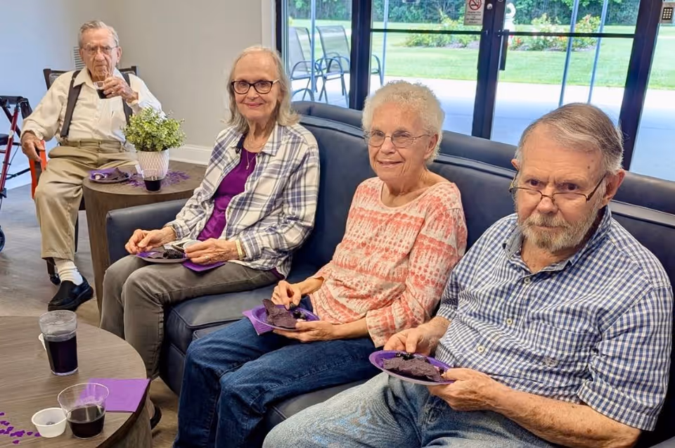 Four older adults sit on couches in a communal lounge eating dessert and drinking from cups.