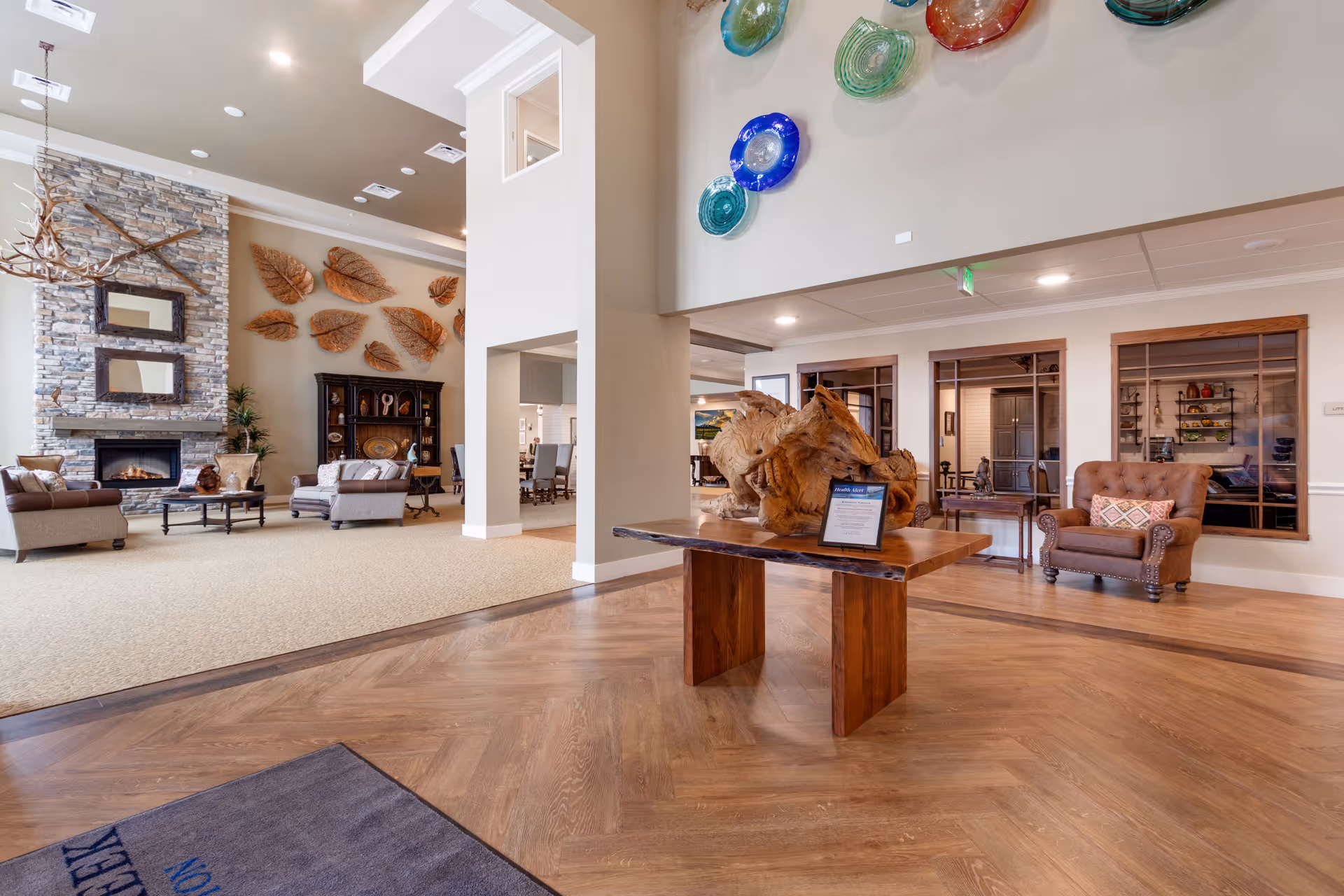 Spacious senior living lobby with seating area, stone fireplace, decorative wall art, and a central wooden table with a sculpture.