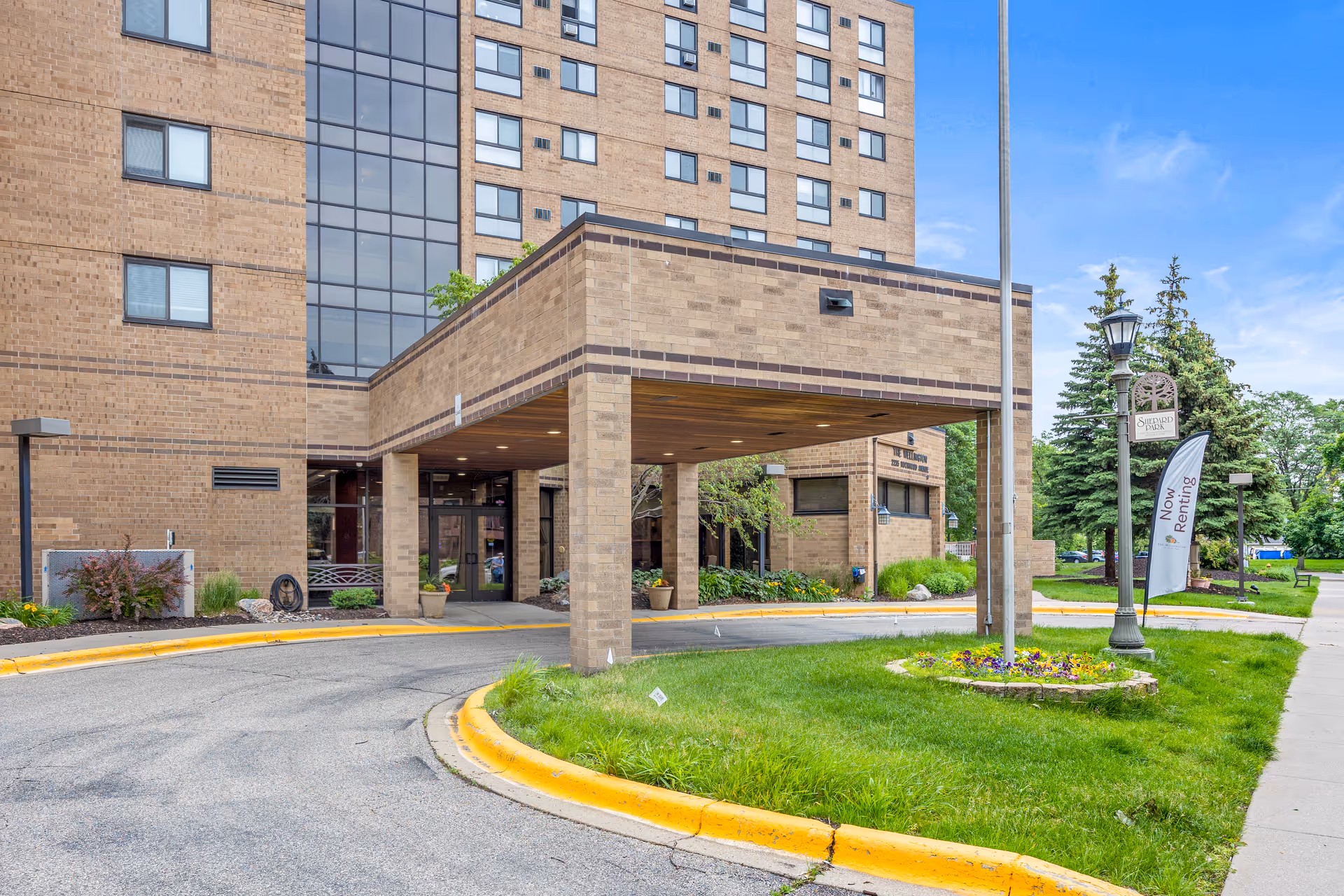 Exterior view of The Wellington Senior Living building entrance with a covered drop-off area, brick facade, a curved driveway, green lawn with flowers, and a lamppost with a sign and a 'Now Renting' flag.