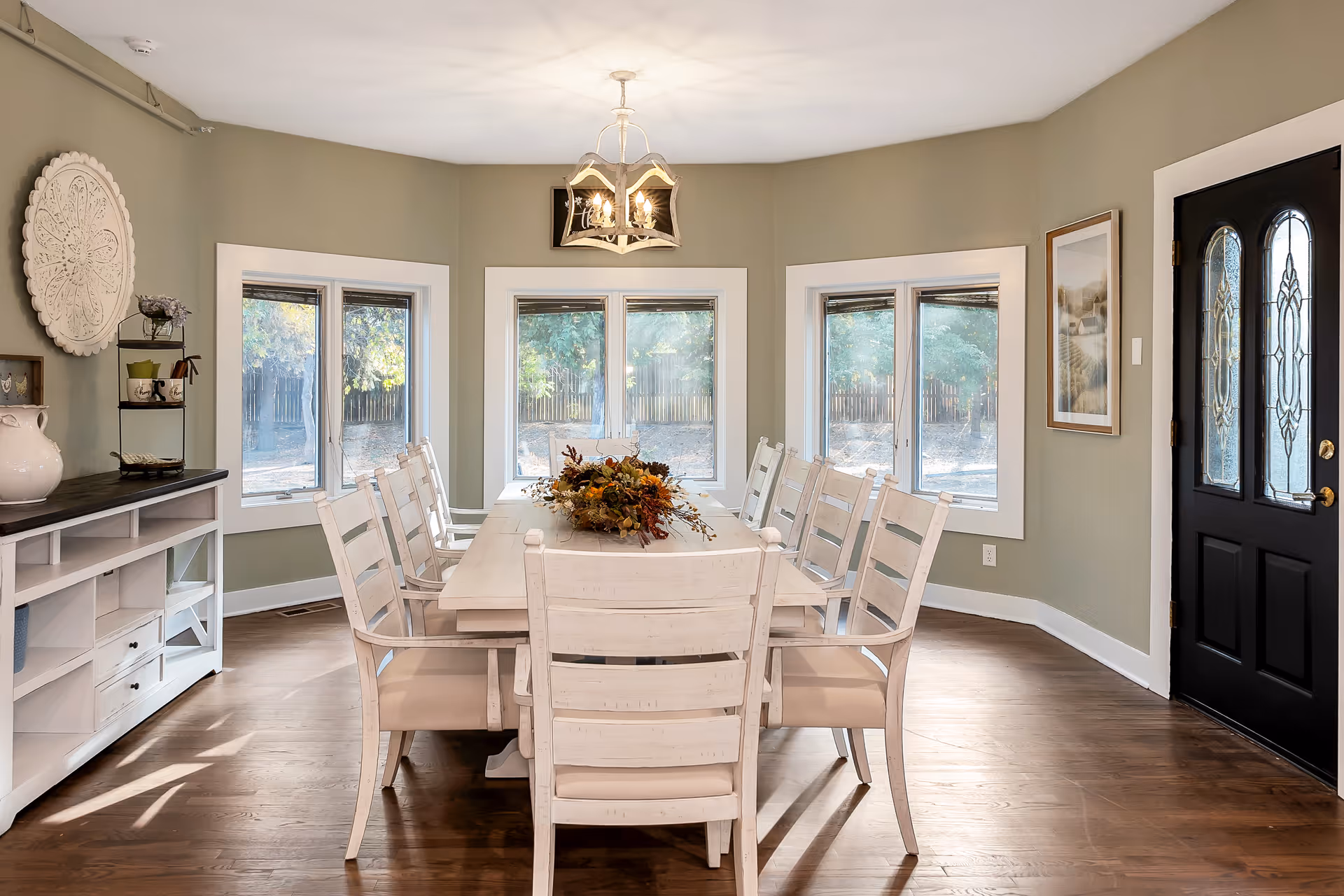 A bright dining room with a long white wooden table surrounded by eight matching chairs. The table is decorated with a centerpiece of autumn foliage. The room has light green walls, three large windows with white trim letting in natural light, a black front door with decorative glass panels, a white sideboard with shelves and drawers, and a decorative wall hanging above it. A chandelier with four lights hangs above the table.