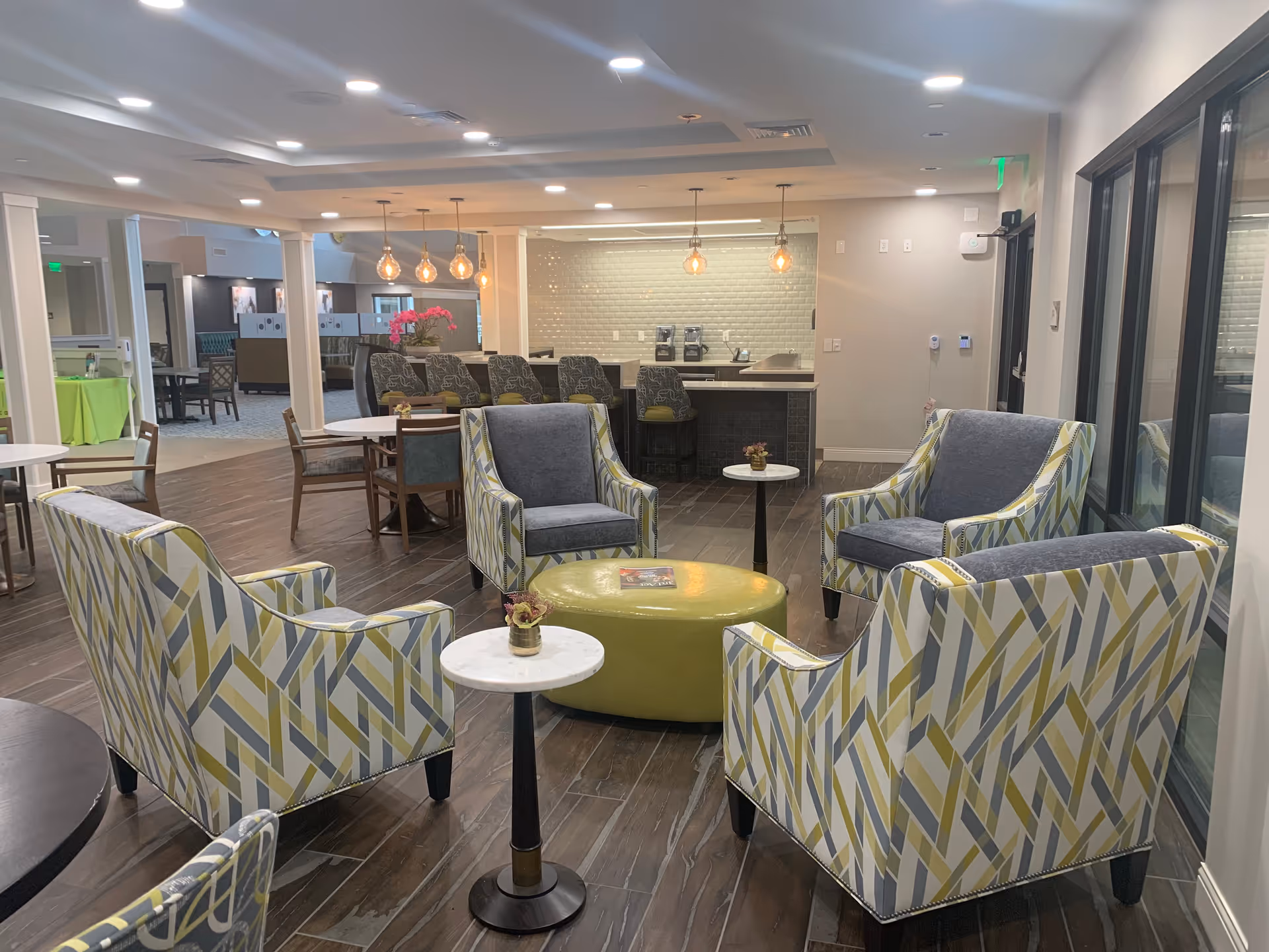 A modern senior living facility common area with patterned armchairs and gray cushioned chairs arranged around a green ottoman and small round tables. In the background, there is a kitchen counter with bar stools and pendant lights hanging from the ceiling. The room has wood flooring and large windows on the right side.