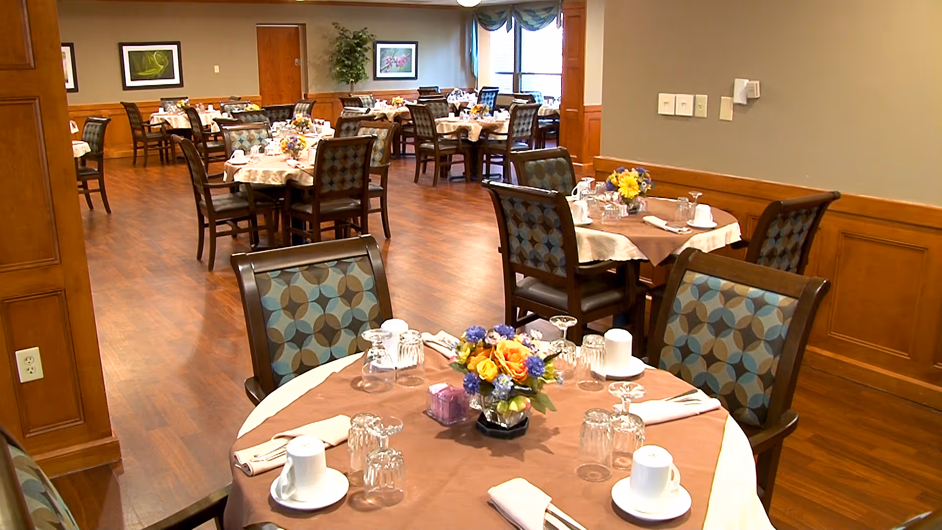 A dining room in a senior living facility with multiple round tables covered with brown and beige tablecloths. Each table is set with cups, glasses, napkins, and floral centerpieces. The room has wooden flooring, patterned chairs, framed artwork on the walls, and a large window with curtains allowing natural light.