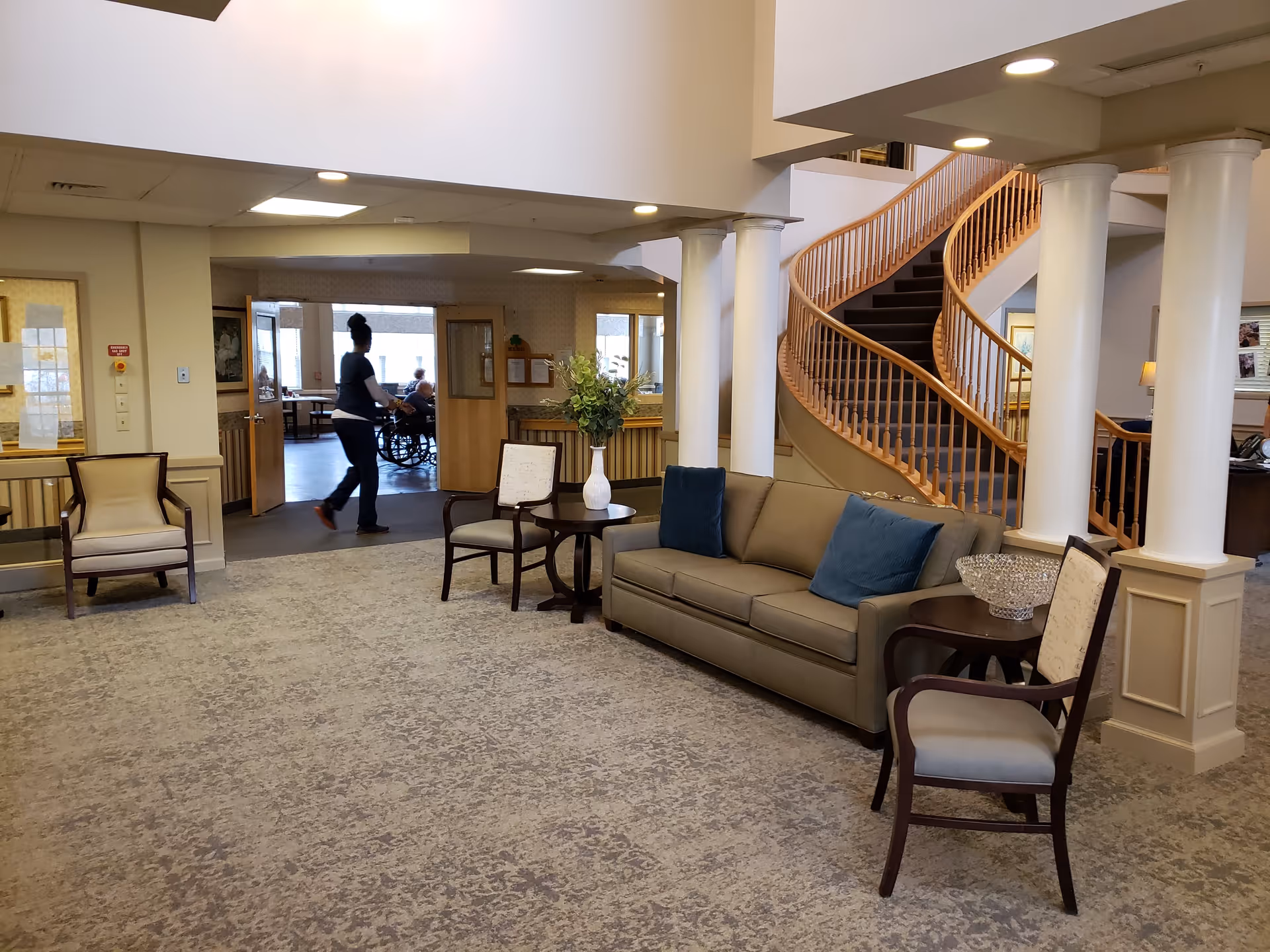 A spacious senior living facility common area with a beige sofa adorned with blue pillows, two armchairs, and a small round table with a vase of flowers. In the background, a person is walking past an open doorway where another person in a wheelchair is visible. A curved wooden staircase with white columns is also visible.