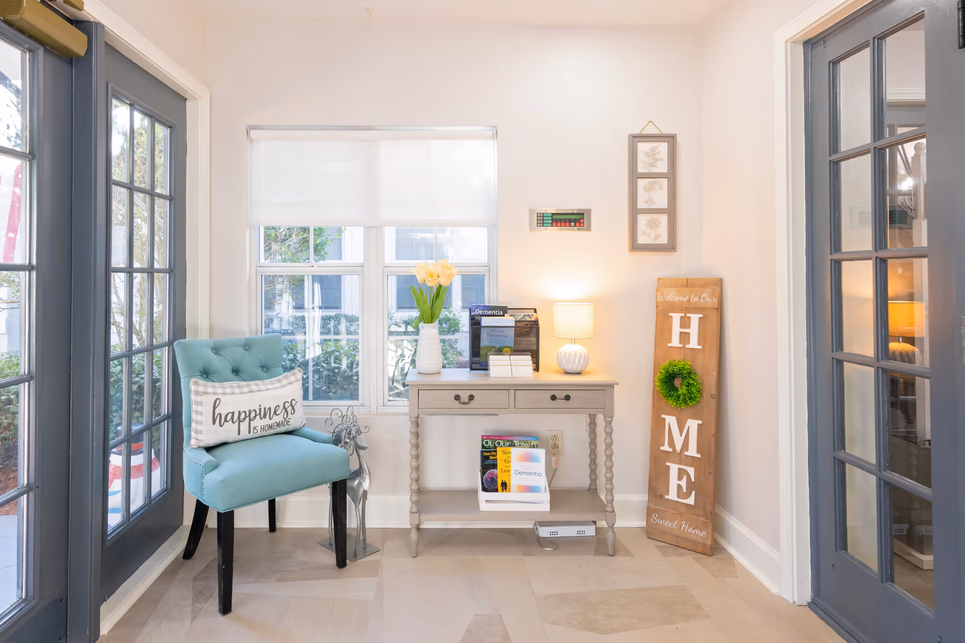 Bright interior seating area with a blue tufted chair, console table with flowers and lamp, and a wooden 'HOME' sign by French doors.