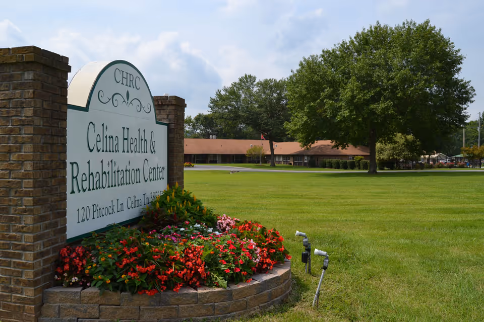 A large green lawn with a flower bed in front of a brick sign that reads 'Celina Health & Rehabilitation Center, 120 Pitcock Ln, Celina TN 38551'. In the background, there is a single-story building with a brown roof and several trees under a partly cloudy sky.