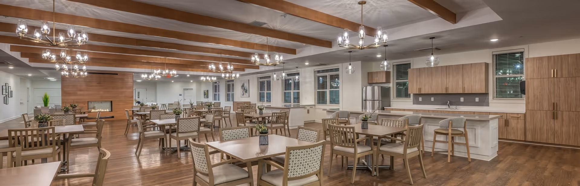 A spacious and well-lit dining area in a senior living facility featuring multiple wooden tables and chairs arranged neatly. The room has wooden ceiling beams, modern chandeliers, and a fireplace with a wooden panel backdrop. There is a kitchen area with wooden cabinets, a refrigerator, and a countertop with bar stools. Potted plants decorate the tables, and large windows with blinds line the walls.