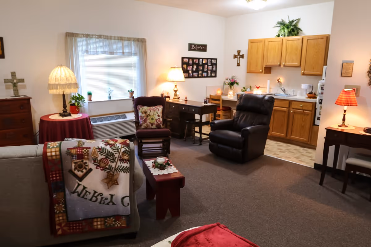 Cozy senior living suite with a sofa, armchairs, small tables and lamps, and a kitchenette with wooden cabinets.