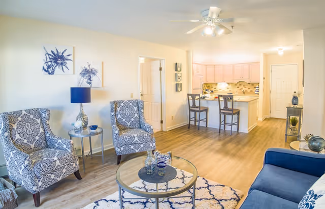 Bright living room with patterned armchairs, a round glass coffee table, and an open kitchen with bar stools in the background.