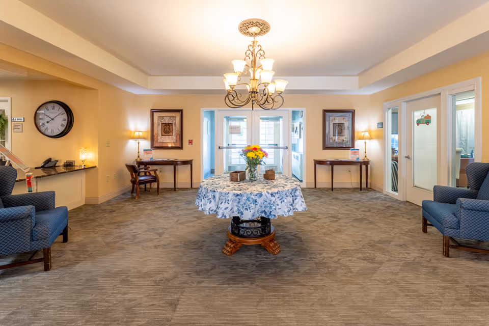 A well-lit common area in a senior living facility with a round table covered by a floral tablecloth and a vase of colorful flowers in the center. Two blue upholstered armchairs are positioned on either side of the room. There are two small wooden tables with lamps and framed artwork on the walls behind them. A large clock is mounted on the left wall above a counter with a phone and other items. Double glass doors are visible at the back of the room, leading to another area.