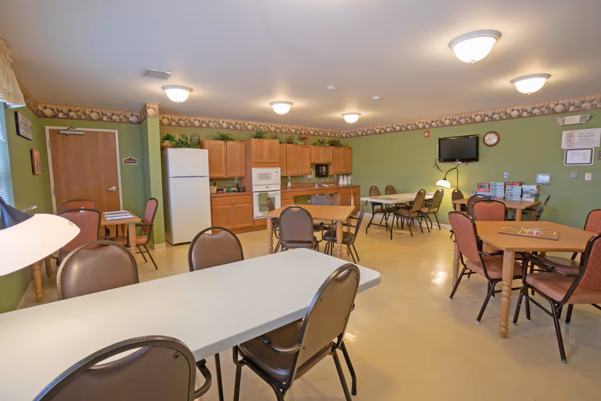 A communal dining and activity room with several tables and chairs arranged throughout. The room has green walls with a decorative border near the ceiling. There is a kitchen area along one wall with wooden cabinets, a white refrigerator, microwave, and oven. A wall-mounted TV and clock are visible on the far wall, along with some board games stacked on a table. The room is well-lit with ceiling lights.