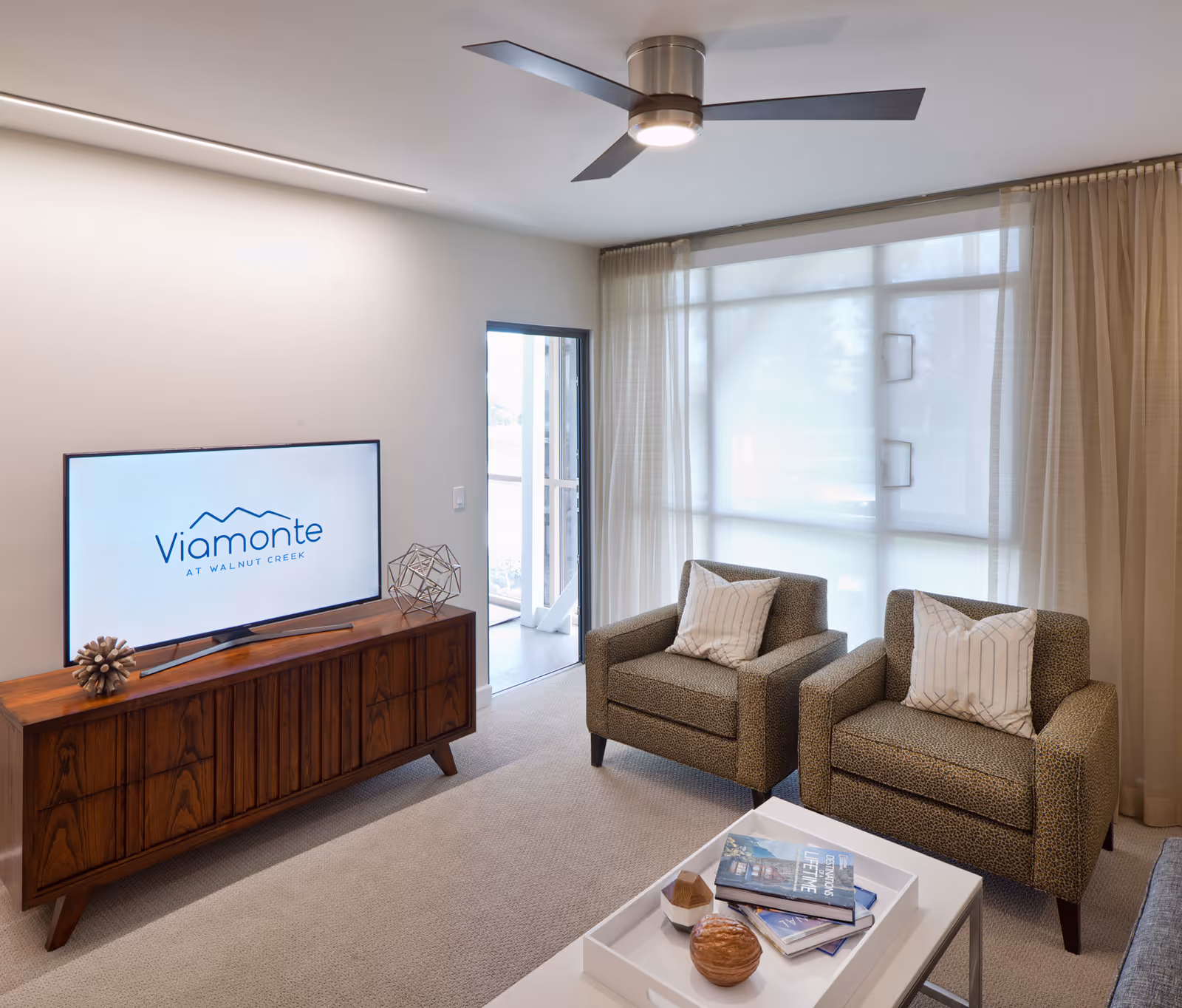 A modern living room with two patterned armchairs each with a white pillow, a wooden TV stand with decorative objects, a flat-screen TV displaying the logo 'Viamonte at Walnut Creek', a white coffee table with books and decorative items, beige carpet, large window with sheer curtains, and a ceiling fan with three blades.