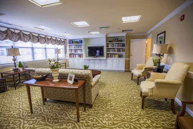 A cozy living room area with patterned carpet, two upholstered armchairs, a sofa, and a wooden table with decorative items. There is a built-in white bookshelf with books and a flat-screen TV mounted in the center. Large windows with valances allow natural light to fill the room.