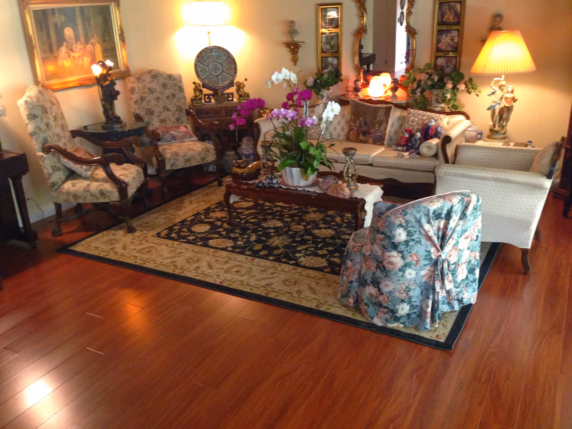 A cozy living room with wooden flooring, a patterned area rug, a floral armchair, two upholstered armchairs, a beige sofa with decorative pillows, a wooden coffee table with potted orchids, and various lamps and decorative items including framed artwork and mirrors on the walls.