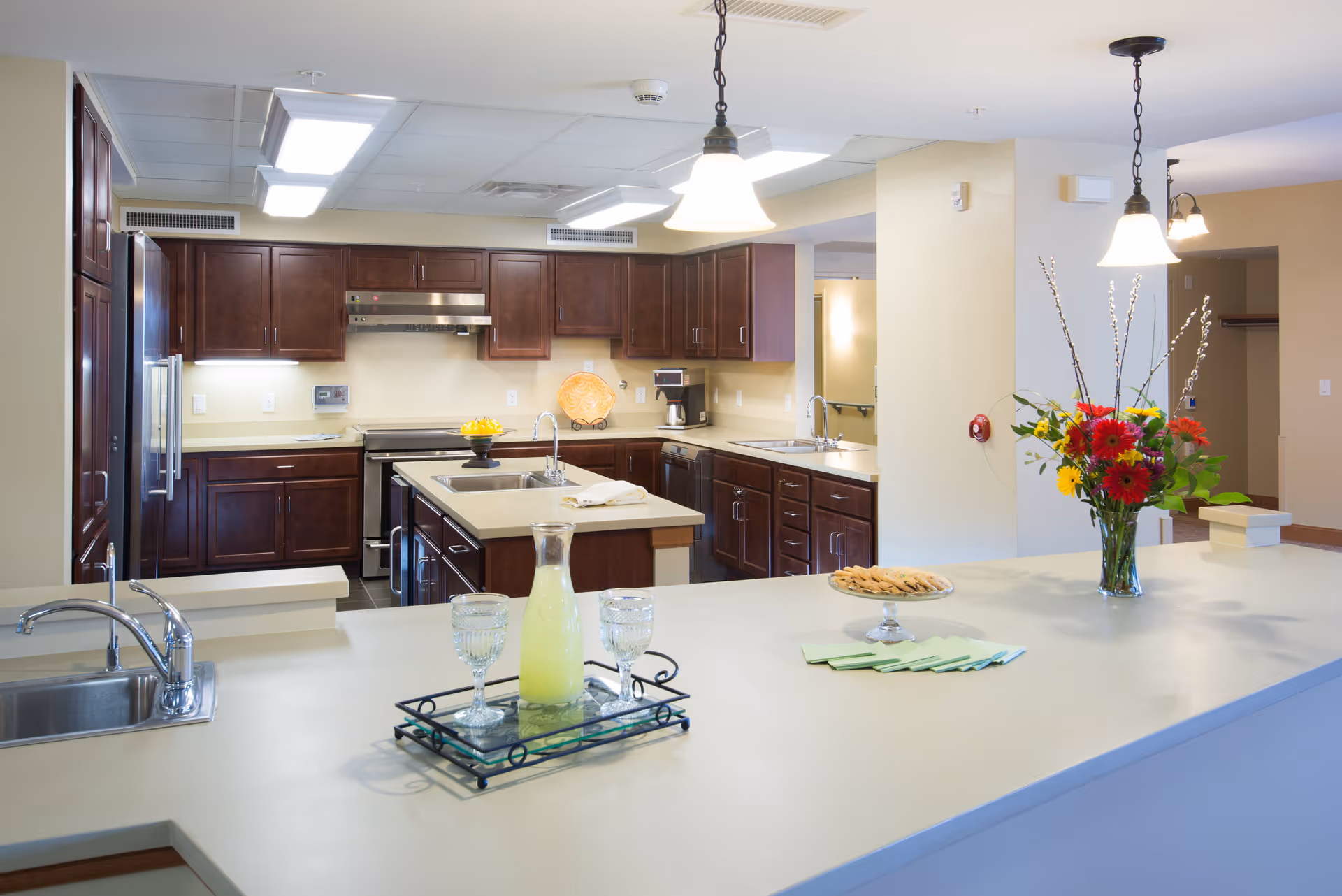 Bright and spacious kitchen with dark wooden cabinets, a large island with a sink, a tray with a pitcher of lemonade and two glasses, a plate of cookies, and a vase of colorful flowers on the counter. The kitchen has modern appliances and pendant lights hanging from the ceiling.