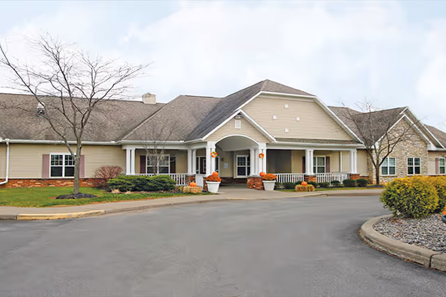Front exterior view of a single-story senior living facility building with beige siding, a covered entrance with white columns, and landscaped areas with small trees and shrubs.
