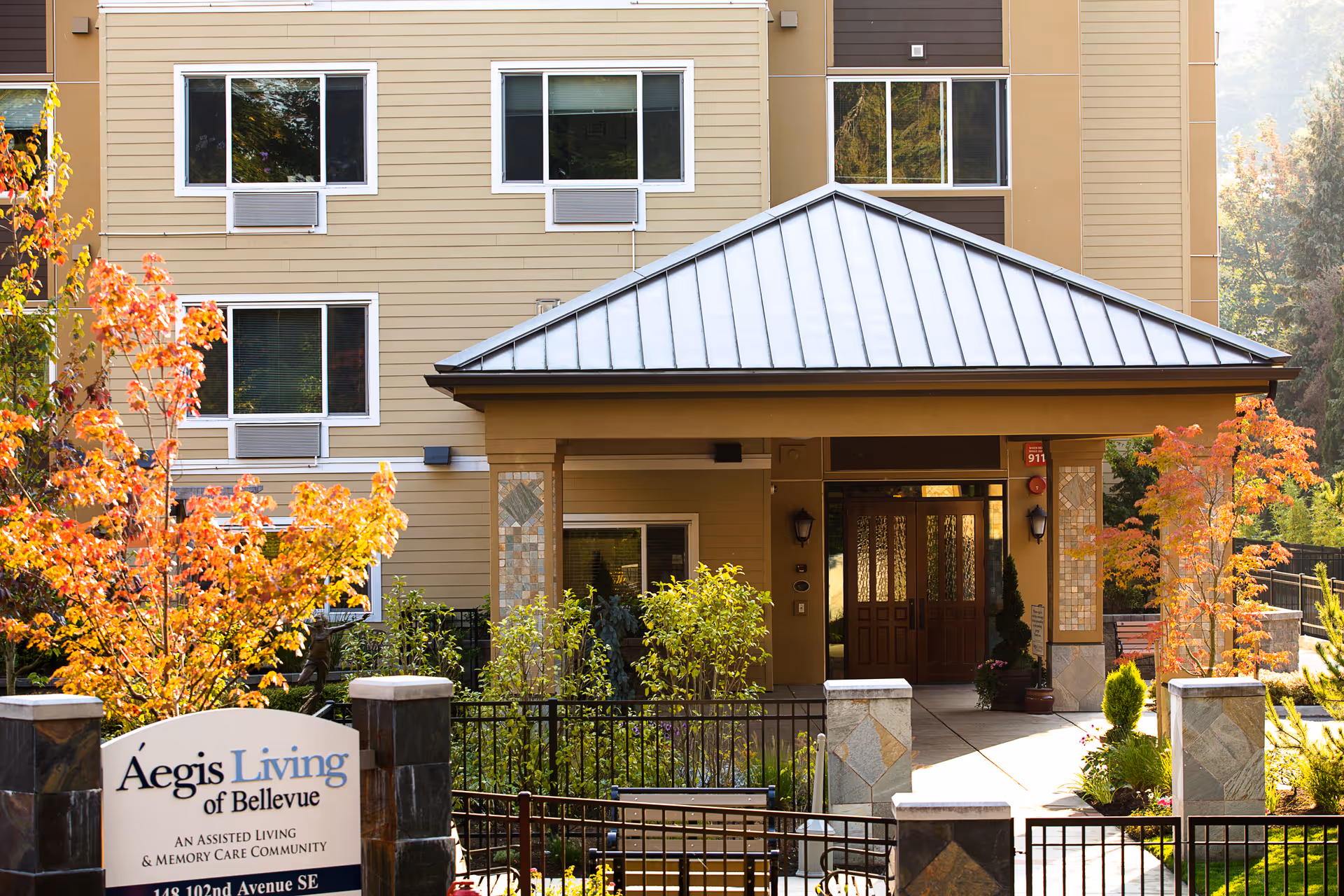 Exterior view of Aegis Living of Bellevue, an assisted living and memory care community, showing the entrance with a covered porch, surrounding greenery, and autumn-colored trees.