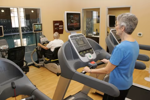 An elderly woman in a blue shirt walking on a treadmill in a fitness room, while an elderly man is using a recumbent exercise bike in the background. The room has light-colored walls, wooden flooring, and several exercise machines with screens. There are windows and mirrors on the walls.