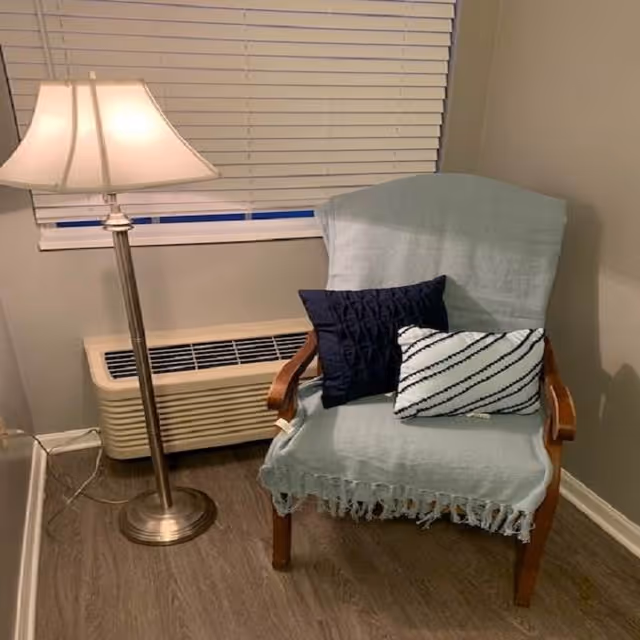 A cozy corner featuring a wooden armchair draped with a light blue throw blanket and two decorative pillows, one navy blue and one white with black diagonal stripes. Next to the chair is a tall floor lamp with a white lampshade, and behind the chair is a window with closed white blinds and a beige air conditioning unit below it. The floor is covered with wood-style flooring.