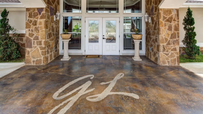 Entrance to a building with double glass doors framed by stone pillars and two decorative planters on pedestals. The ground in front of the entrance has a large painted monogram with the letters 'GL'.