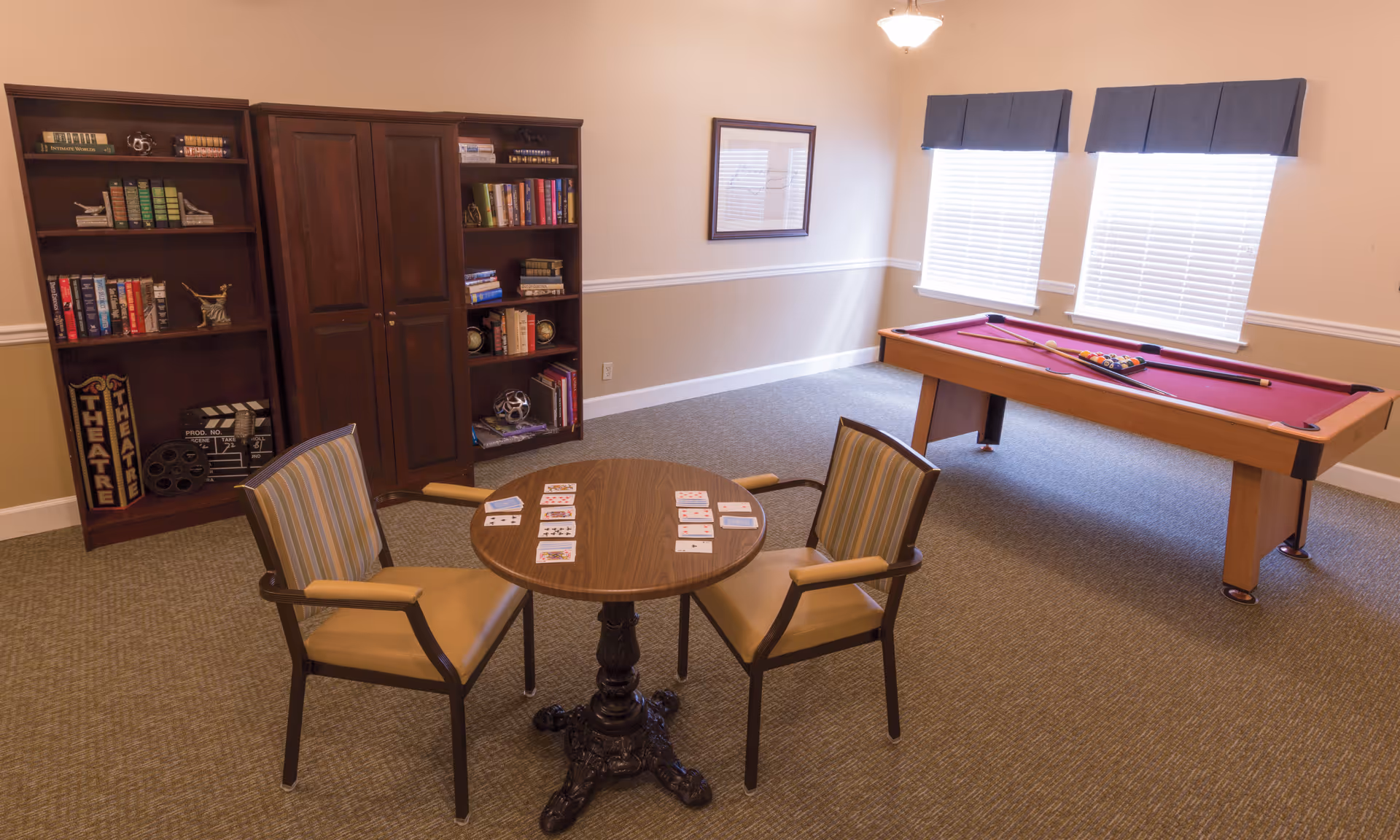A senior living community game room with a pool table, a round card table with two chairs, and bookshelves against the wall.